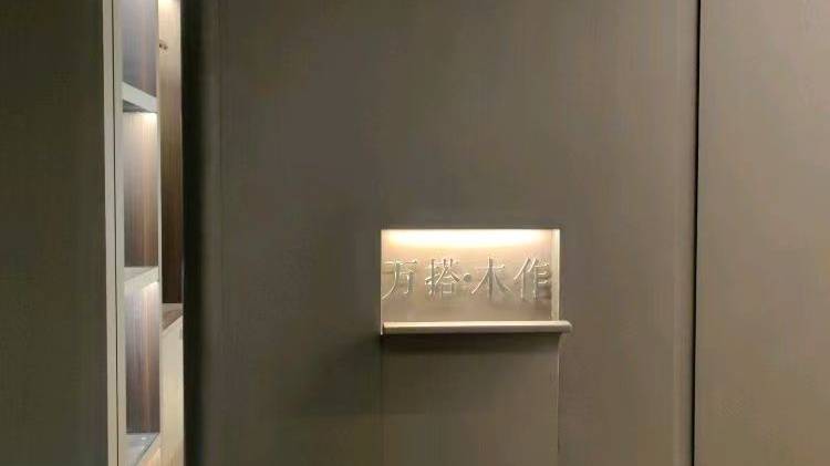 这是一张家居建材展厅或全屋定制门店的实拍图。画面主体为深灰色哑光饰面墙体，中央设有内嵌式发光标识槽，清晰展示“万搭·木作”品牌字样，散发柔和暖光。左侧可见局部开
